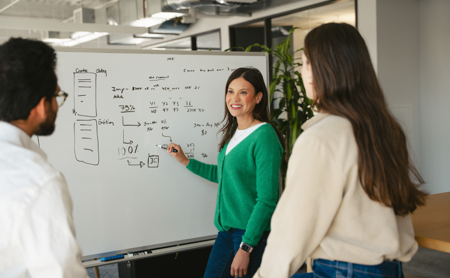 Three people stand around a whiteboard in an office. One woman, holding a marker and smiling, explains diagrams and notes written on the board to her two colleagues.
