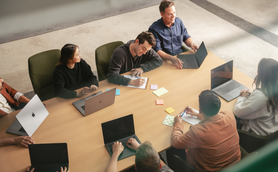 A group of eight people sits around a conference table with laptops, notebooks, and colorful sticky notes, engaged in a meeting or discussion in a modern office setting.
