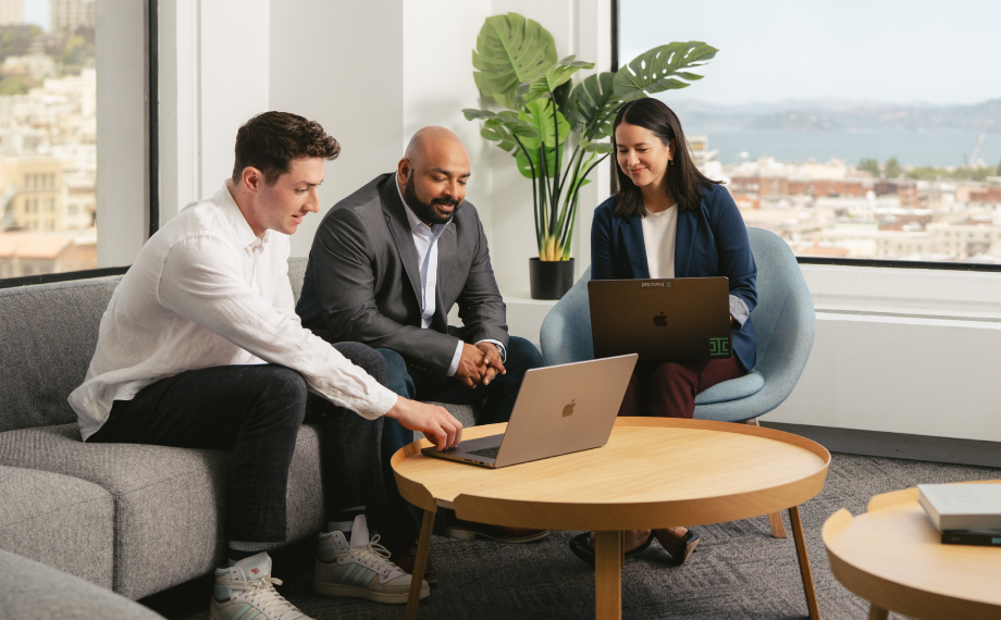 Three people sit around a wooden coffee table in a modern office with large windows, two using laptops and one pointing at a screen, collaborating in a relaxed, professional setting with a cityscape view in the background.
