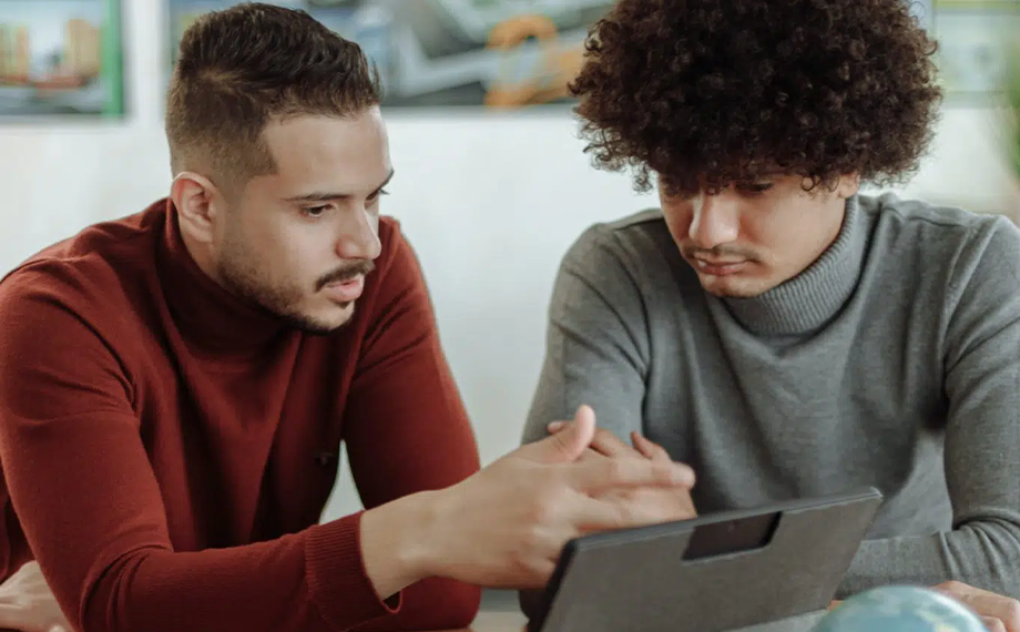 Two young men sit side by side at a table, looking intently at a tablet screen. As part of a negotiation process, one in a red turtleneck gestures while the other in a gray sweater listens attentively.