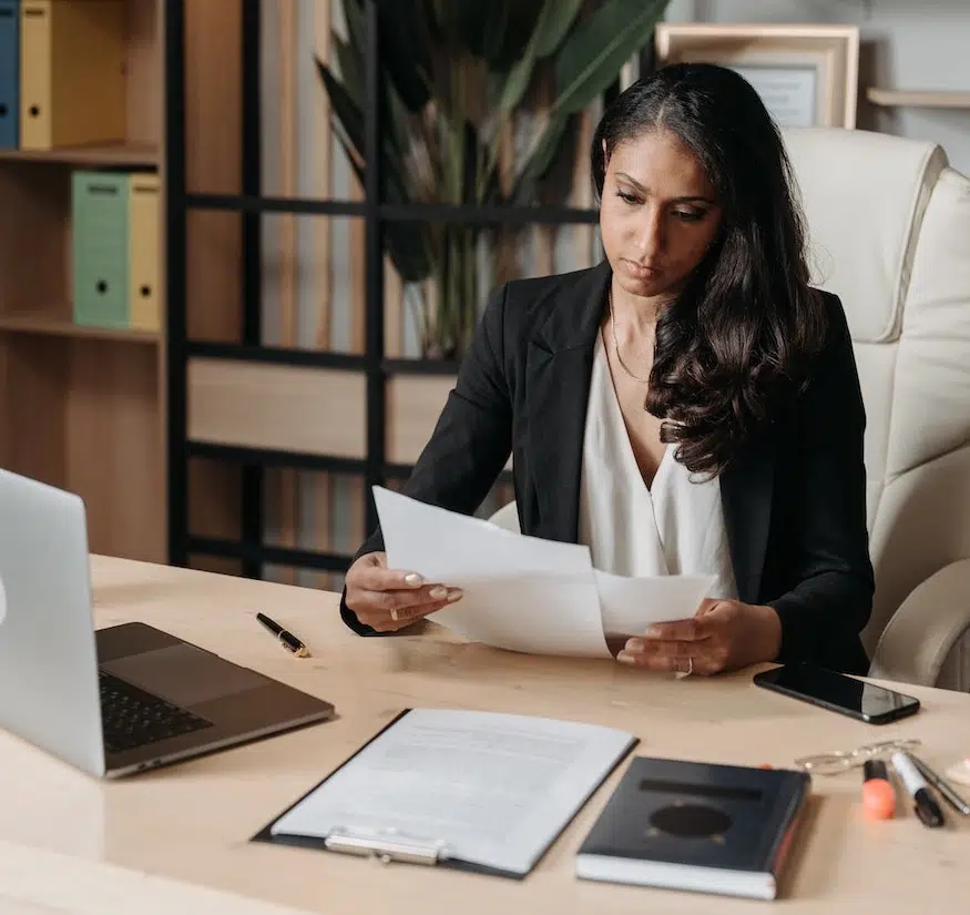 A woman in business attire sits at a desk reviewing contract amendments, with a laptop, notebook, smartphone, and office supplies in front of her. Shelves and plants are visible in the background.