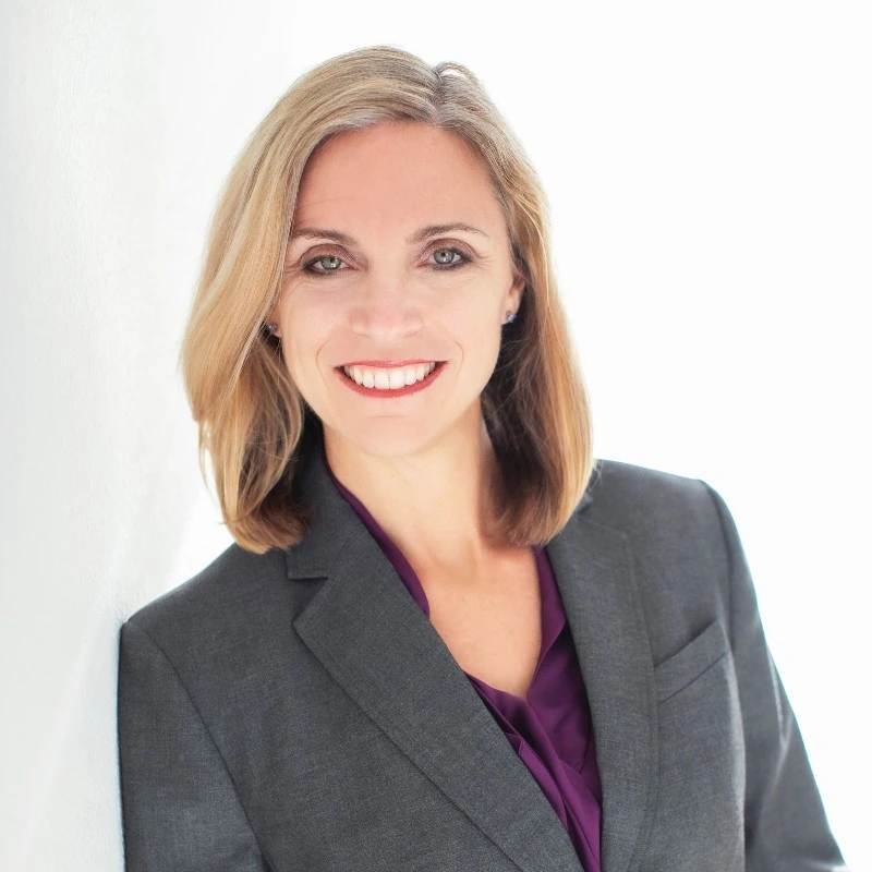 A woman with shoulder-length blonde hair, wearing a dark gray blazer over a purple blouse, smiles at the camera against a bright, white background, representing a modern juro alternative.