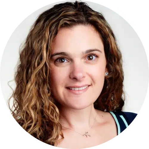 A woman with long, curly brown hair and light skin smiles at the camera. She is wearing a dark top with light trim and a delicate necklace, posed against a plain, light background, perfect for a docusign alternative business profile.