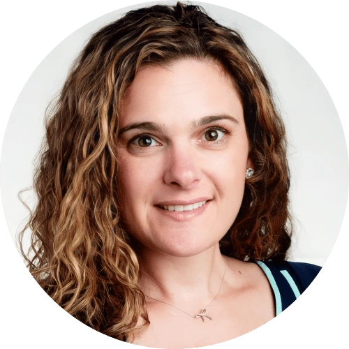 A woman with long curly brown hair, wearing a necklace and a dark top with white trim, smiles at the camera against a plain light background, showcasing her unique style—a true juro alternative vibe.