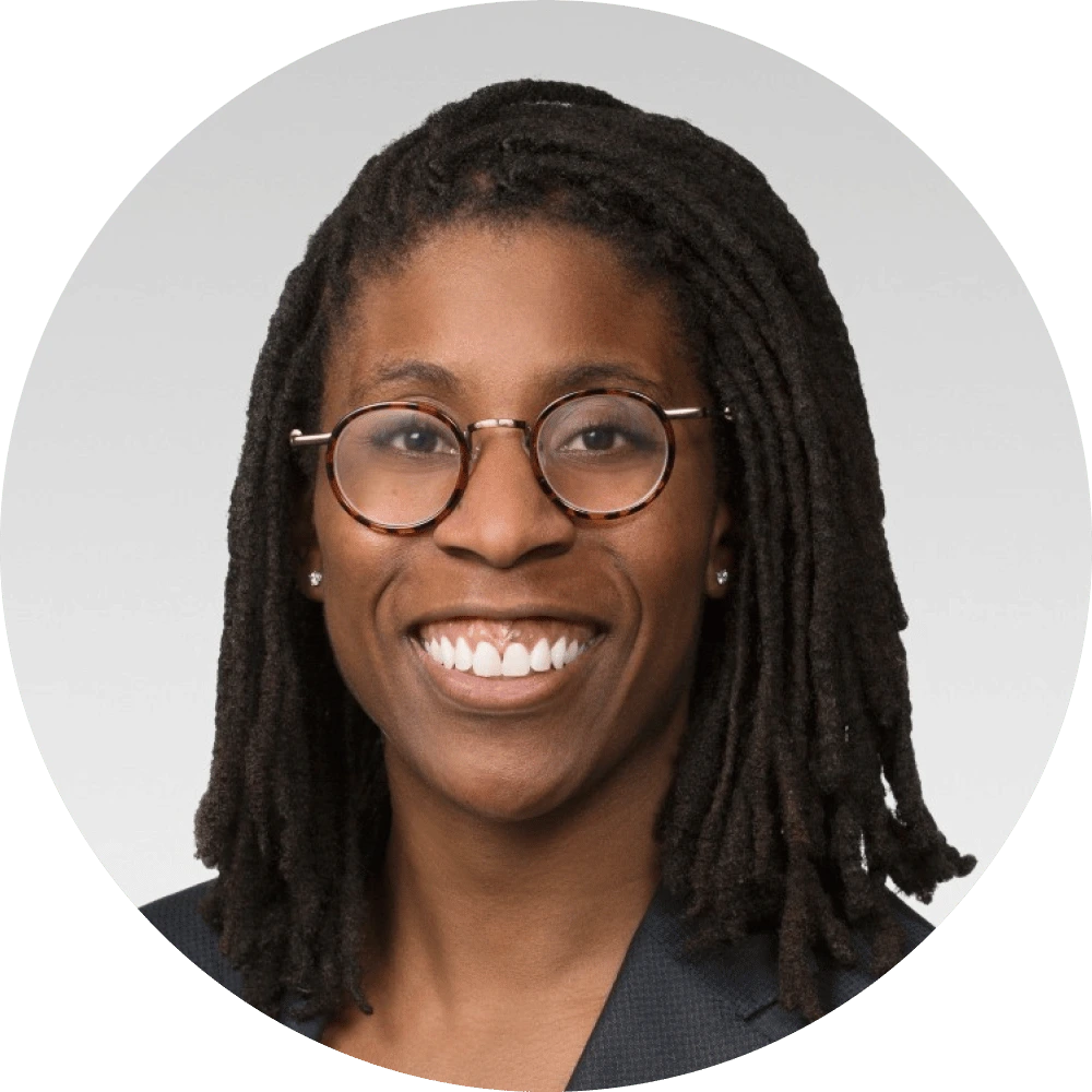A person with medium-length dreadlocks, wearing round glasses and a dark blazer, smiles at the camera against a light gray background—perfect for showcasing an icertis alternative professional team member.