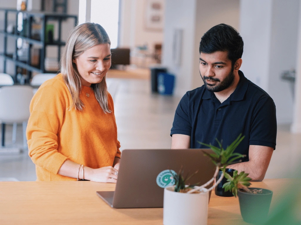 Two people, one wearing an orange sweater and the other in a dark polo shirt, are smiling and looking at a laptop on a table in a bright, modern office with plants.
