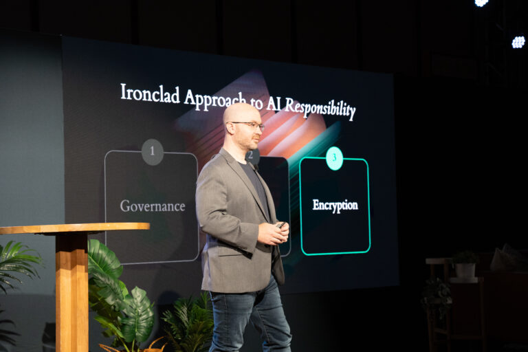 A man in a gray jacket stands on stage giving a presentation about AI responsibility, with a slide behind him displaying the words Governance and Encryption under the title Ironclad Approach to AI Responsibility.