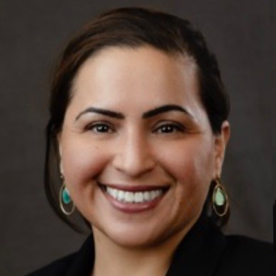 A woman with dark hair pulled back, wearing turquoise drop earrings and a black top, smiles at the camera in front of a plain dark background.