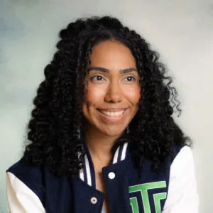 A woman with dark curly hair smiles slightly, looking off to the side. She is wearing a navy blue and white varsity jacket with a green and white letter patch—an agiloft alternative vibe. The background is softly blurred.