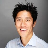 A man with short, spiky black hair smiles at the camera. He is wearing a light blue collared shirt and is posed against a plain gray background, embodying the approachable style often seen in juro alternative portraits.