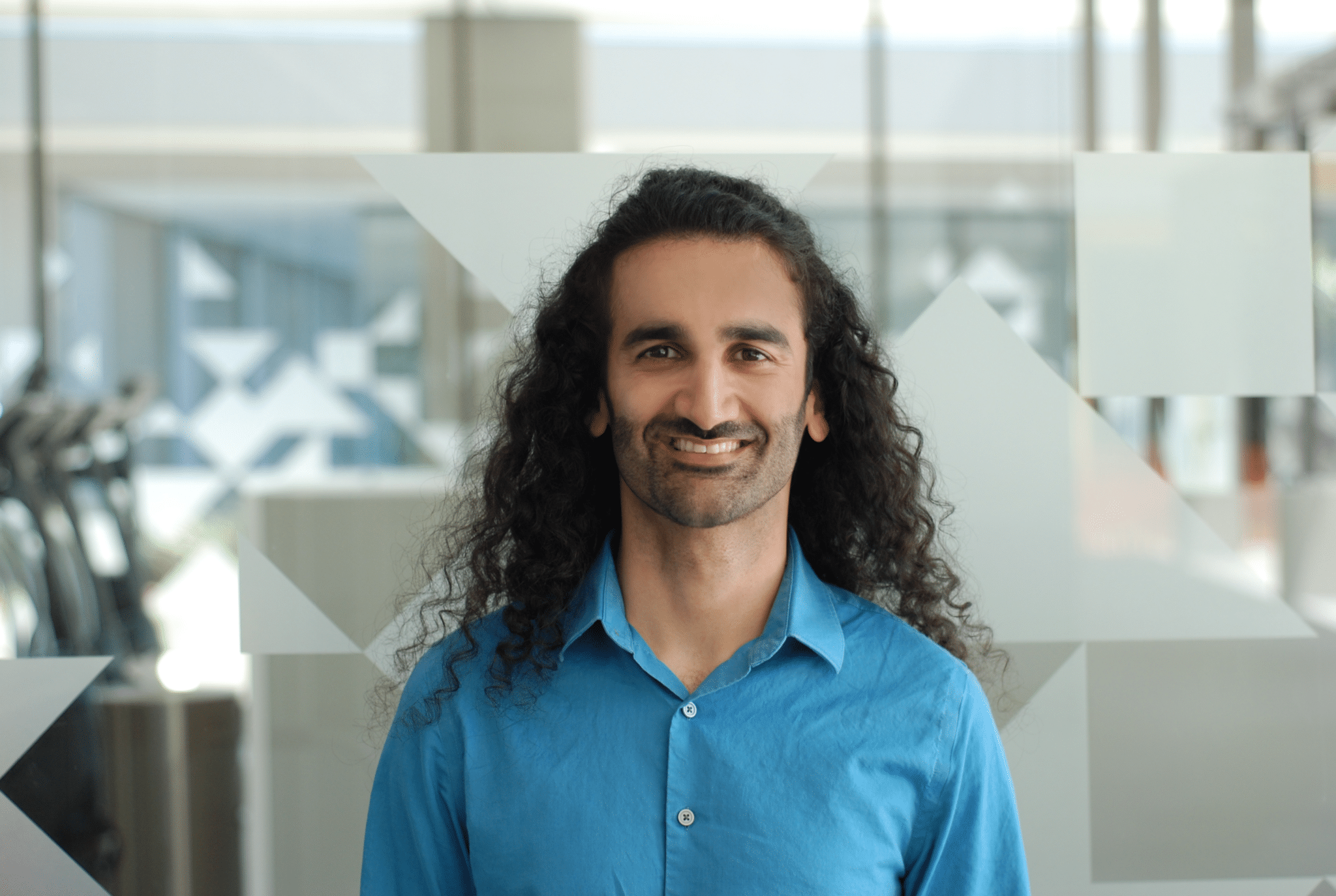 A person with long, curly hair and a beard, wearing a bright blue shirt, smiles while standing indoors in front of a frosted glass wall with geometric patterns.