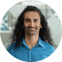 A person with long, curly dark hair and a beard smiles while wearing a blue collared shirt, standing indoors with a blurred, modern background—perfect for representing a juro alternative.