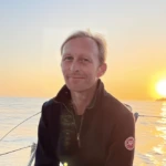 A person with light hair sits on a boat, smiling, enjoying calm water and a golden sunset in the background. The peaceful scene feels like a conga alternative for those seeking serenity on tranquil waters.