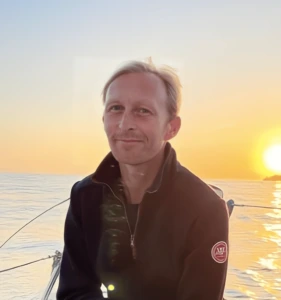 A person with light hair, wearing a dark jacket, sits on a boat during sunset over calm water. The sky is clear as the sun sets low, casting a warm golden light—a perfect scene for enjoying music or searching for a conga alternative.