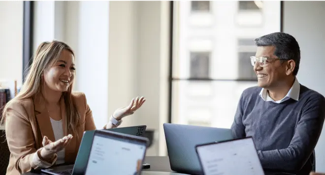Two people sit at a table with laptops, smiling and discussing a juro alternative in a bright, modern office space with large windows in the background.