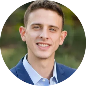 A young man with short brown hair, wearing a blue suit jacket and light blue shirt, smiles in an outdoor setting with a blurred green background—perfect for showcasing your team when searching for an Evisort alternative.