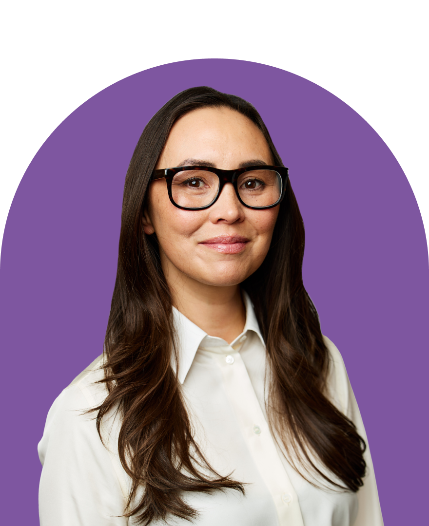 A woman with long brown hair and glasses, wearing a white button-up shirt, smiles slightly in front of a purple semicircular background.