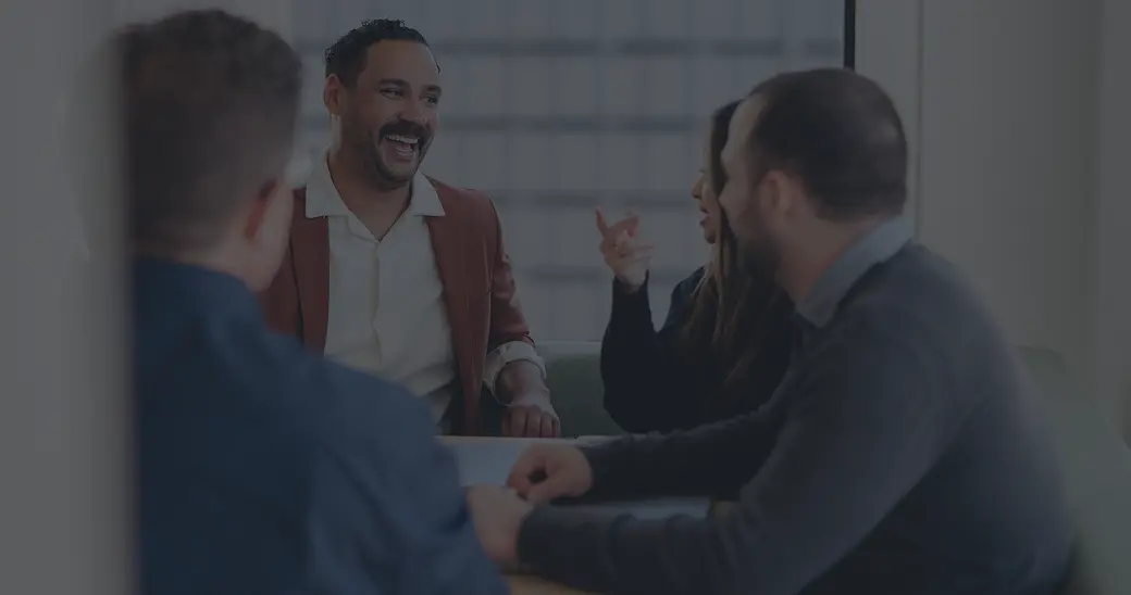Four people sit around a table in an office, engaged in conversation. One man wearing a brown blazer is smiling and laughing, while another person gestures with their hand. The atmosphere appears friendly and collaborative.