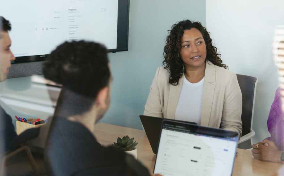 A woman in a beige blazer sits at a conference table with three colleagues, engaged in a meeting discussing ai for career growth. Laptops are open on the table and a screen is visible in the background.