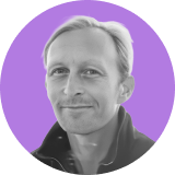 Black and white portrait of a smiling man with light hair, wearing a jacket, shown against a circular purple background, representing the team behind Ironclad CLM.