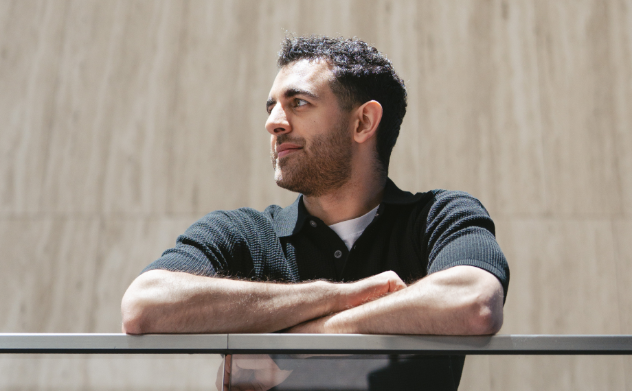 A man with short curly hair and a trimmed beard leans on a railing, looking to the side with a slight smile, as if contemplating how ai for career growth could shape his future. He wears a black polo shirt and stands in front of a plain, beige background.