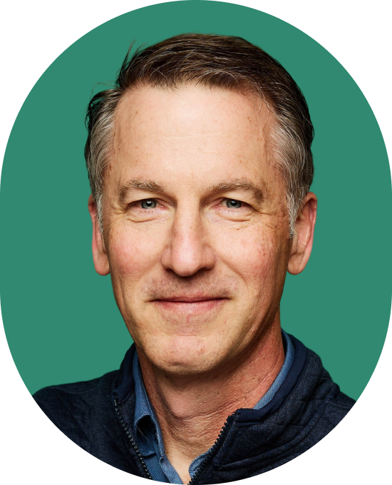 A middle-aged man with short, light brown hair and blue eyes smiles gently. He is wearing a blue collared shirt and a dark jacket, with a green background behind him.