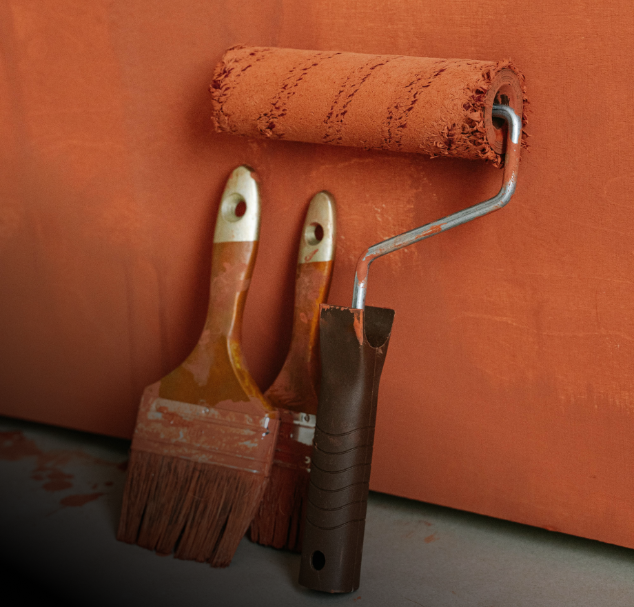 A worn paint roller and two paintbrushes with brown handles rest against a wall painted in a rusty orange color, evoking the careful touch of those who create contracts between craftsmanship and creativity. Some paint remains on the brushes and roller.