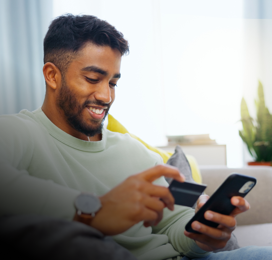 A smiling man sits on a couch, holding a credit card in one hand and a smartphone in the other, appearing to make an online purchase or create contracts. Cushions and a plant decorate the background.